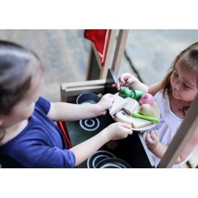 Crianças a brincar numa cozinha de brinquedo de madeira com utensílios e alimentos de plástico coloridos.