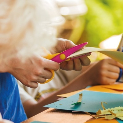 Tesoura amarela e rosa a cortar papel colorido numa mesa com folhas e pessoas ao fundo