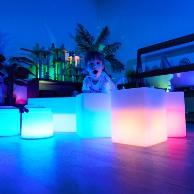 Criança com cubos luminosos coloridos em ambiente interior com plantas e estantes.
