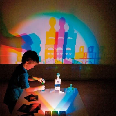 Criança a brincar com peças coloridas numa mesa com projeções de sombras coloridas na parede