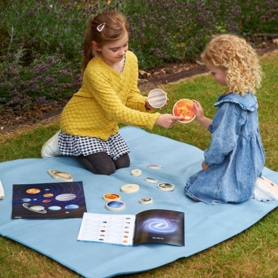Duas crianças brincando com peças do sistema solar num tapete azul na relva