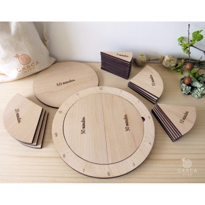 Relógio didático de madeira com segmentos destacáveis marcados com unidades de tempo em minutos, sobre mesa de madeira com saco ao fundo