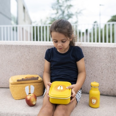 Criança com lancheira, mochila térmica e garrafa amarelas com padrão e cara de urso sentada num banco exterior