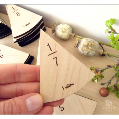 Peça triangular de madeira clara com texto '1/7' e '1 sétima' em fundo com outras peças de madeira e ramo seco.