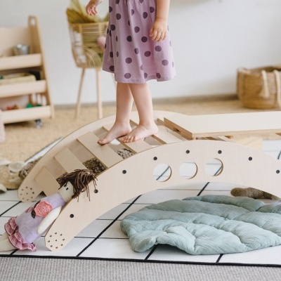 Menina em vestido lilás com bolinhas a pisar em brinquedo de madeira arqueado com boneca de pano e almofada.