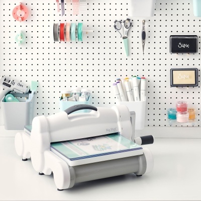 Máquina de corte branca Sizzix Big Shot em mesa com ferramentas de artesanato e painel perfurado ao fundo
