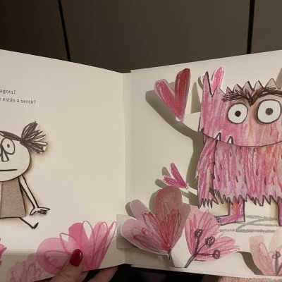 Livro infantil com ilustrações brilhantes de uma menina e um monstro rosa entre flores.