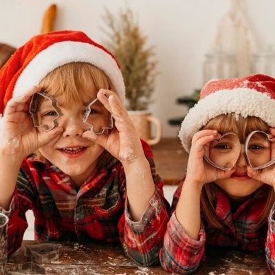 Duas crianças com chapéus de Pai Natal seguram formas de metal para biscoitos à frente dos olhos numa cozinha