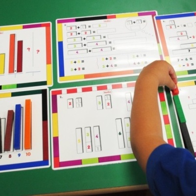 Folhas com exercícios de matemática coloridos sobre mesa verde com mão de criança interagindo