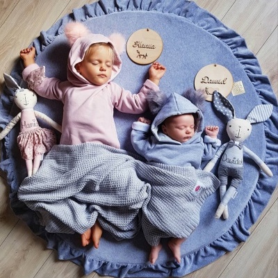 Dois bebés com roupas azuis e rosas a dormir numa manta azul redonda com folhos, rodeados por bonecas de pano e placas com nomes.