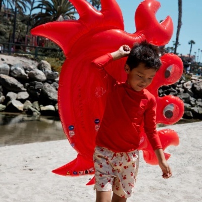 Criança na praia com bóia insuflável vermelha em forma de caranguejo.