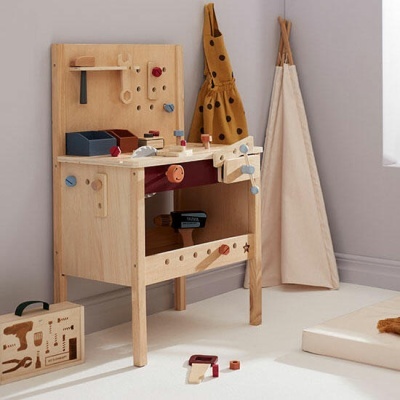 Bancada de trabalho infantil de madeira com ferramentas de brinquedo, mochila castanha com pontos pretos e tenda de tecido bege num quarto claro.