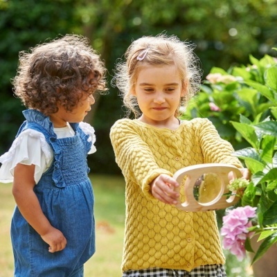 Duas crianças ao ar livre, uma com cardigan amarelo segurando uma lente de aumento perto de flores rosas, outra com macacão azul.