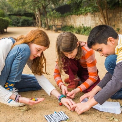 Crianças ao ar livre a jogar com pequenos objetos e tabuleiro quadriculado.
