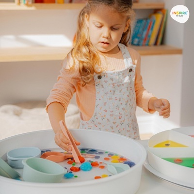 Criança loira a brincar com bandeja de bolas coloridas numa mesa branca