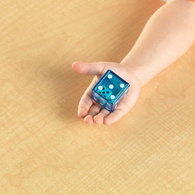 Mão infantil segurando dado azul translúcido com pontos brancos sobre mesa de madeira clara