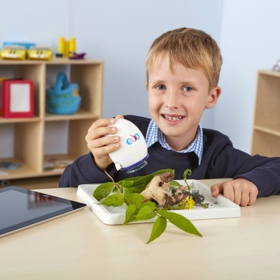 Criança despejando líquido sobre plantas num tabuleiro branco numa mesa clara com um tablet ao lado