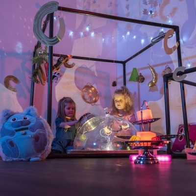 Crianças brincando dentro de estrutura cúbica decorada com balões e luzes, bolo artificial iluminado e peluche azul.