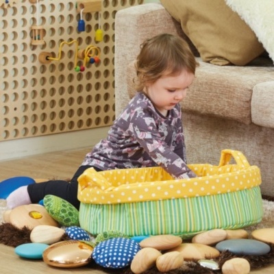 Criança a brincar com almofadas coloridas em cesta de tecido amarela no chão junto a sofá e painel lúdico no quarto