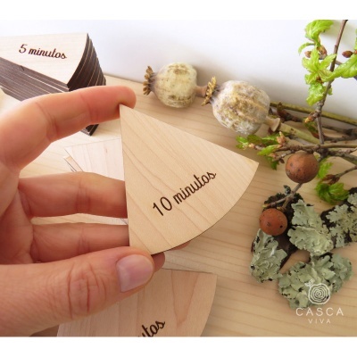 Peça de madeira em forma de fatia com inscrição '10 minutos' sendo segurada por mão, com elementos naturais ao lado.