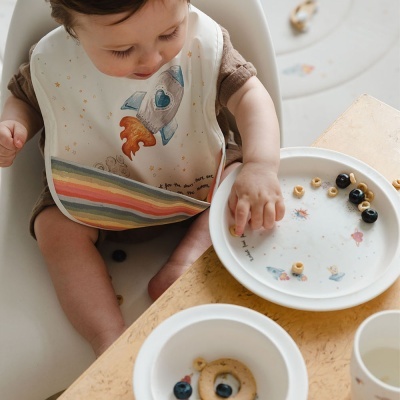 Bebé com babete estampado com foguete e arco-íris a comer cereais em pratos brancos sobre mesa de madeira