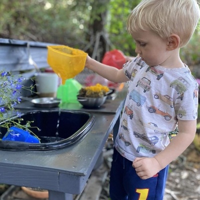 Criança a despejar água de jarra amarela numa cuba preta numa mesinha cinzenta ao ar livre com plantas