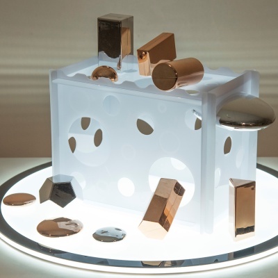 Caixa branca translúcida com peças metálicas douradas e prateadas ao redor sobre base iluminada