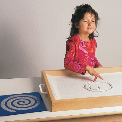 Criança a desenhar espiral em caixa de areia com tabuleiro de madeira e tapete azul
