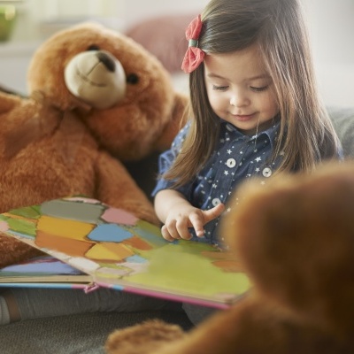 Criança pequena a ler livro colorido sentada num sofá com dois ursinhos de peluche castanhos maiores