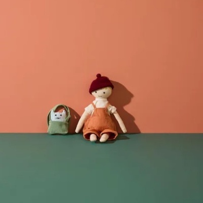 Boneco de pano com macacão laranja e gorro vermelho ao lado de mochila verde sobre superfície verde e fundo salmão.