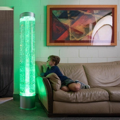 Coluna luminosa verde com bolhas ao lado de sofá de pele bege com menino sentado, parede branca de tijolos e quadro abstrato