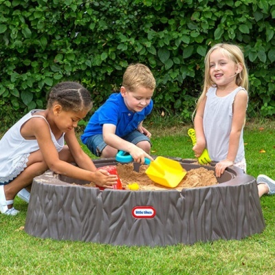 Caixa de areia infantil castanha com três crianças a brincar com pás e baldes no relvado