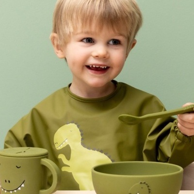 Copo, t-shirt e tigela verdes com dinossauros, criança sorrindo sentada a uma mesa