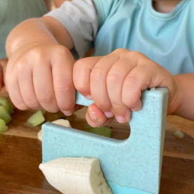 Mãos de criança usando cortador plástico azul claro para cortar banana sobre tábua de madeira