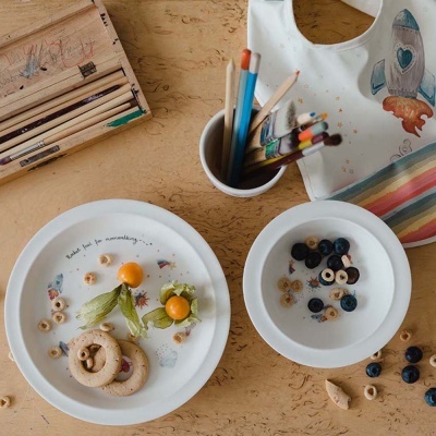 Conjunto de louça branca com alimentos, lápis de cor e babete estampado sobre mesa de madeira