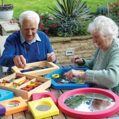 Dois idosos jogam com tabuleiros e peças coloridas ao ar livre num banco de madeira