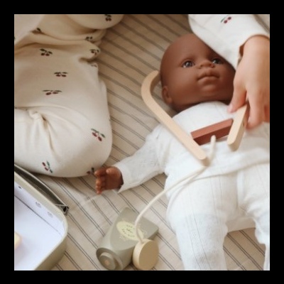 Boneca bebê castanho escuro com roupa branca deitada em colchão riscas cinza e branco com criança e brinquedo de madeira