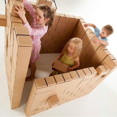 Três crianças brincando numa casa feita de caixas de cartão castanho empilhadas.