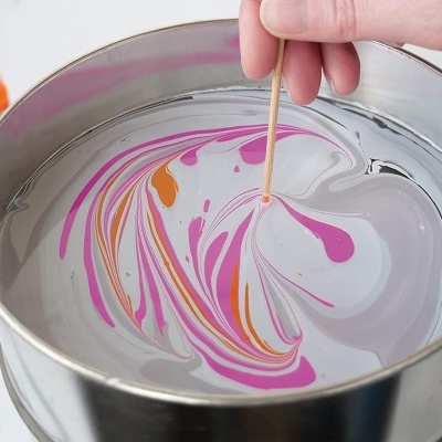 Balde metálico com água e tinta colorida formando padrão marmoreado e mão usando palito para misturar