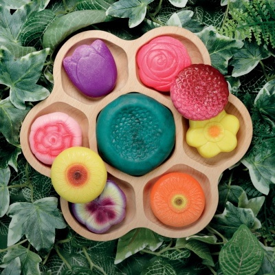 Sabonetes coloridos em forma de flores e frutas em suporte de madeira entre folhas verdes