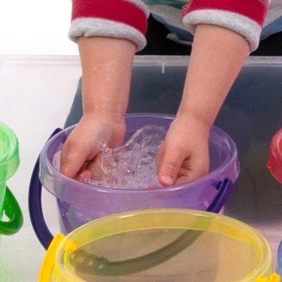 Balde transparente roxo com mãos de criança e água, rodeado por baldes coloridos.