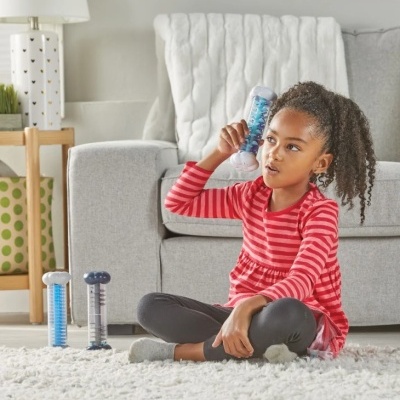 Menina com camisola vermelha segurando brinquedo cilíndrico azul, sentada no chão junto a sofá cinzento