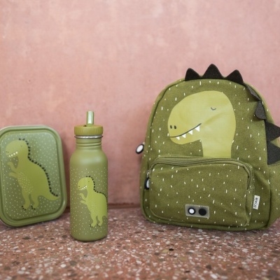 Mochila, garrafa e estojo verdes com dinossauro e detalhes a imitar espinhos, chão pedra, fundo rosa.