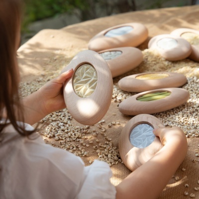 Objetos ovais de madeira com janelas coloridas e texturizadas, criança toca neles sobre superfície com grãos