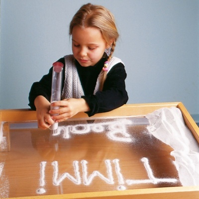 Criança a brincar com areia branca numa caixa de madeira e vidro transparente