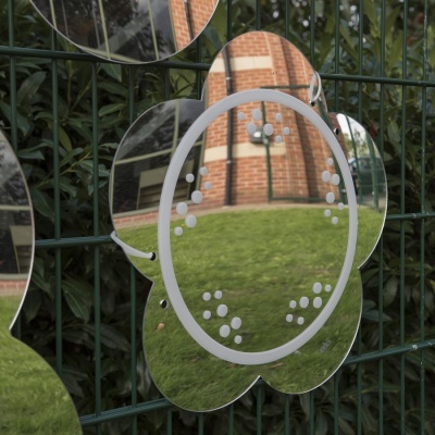 Conjunto de espelhos decorativos em forma de flores numa grade metálica preta com fundo de plantas e edifício de tijolos