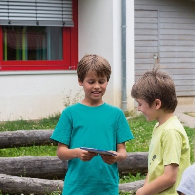 Três crianças no exterior com uma casa ao fundo, uma segura um dispositivo eletrónico.
