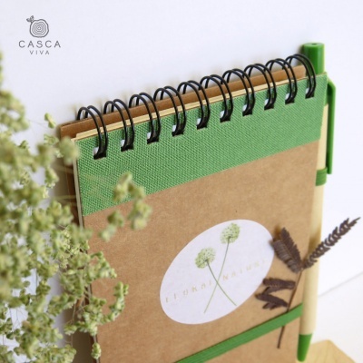 Caderno espiral com capa kraft e verde, caneta de madeira verde, etiqueta com plantas e texto FIDALGA NATURAL