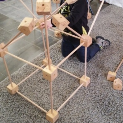 Brinquedo de construção em madeira com cubos e bastões numa sala com tapete cinzento.