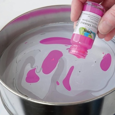 Frasco de tinta cor-de-rosa sendo despejado em bacia metálica com tinta na água e vários frascos de tinta ao fundo.